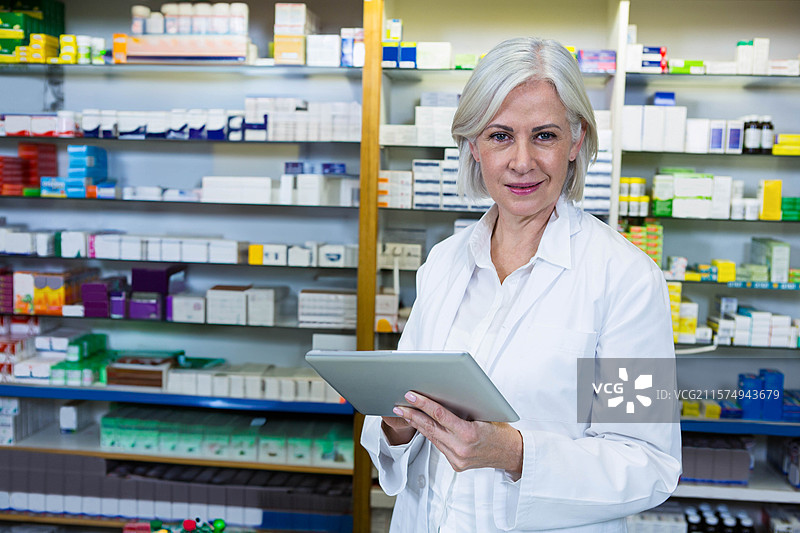 一位资深女性药剂师手持药片，正在药房货架上整理药物，留有空白空间。图片素材