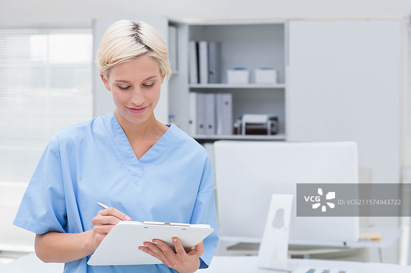 穿着蓝色护士服的护士在诊室里用白色夹板写字，旁边有电脑显示器和键盘。图片素材