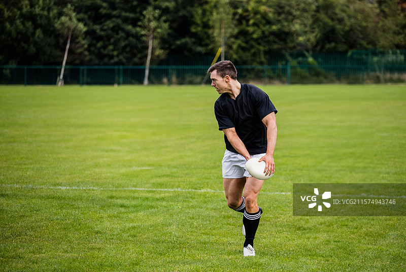 身穿运动服的男性运动员站在草地上，手持白色橄榄球，旁边有围栏，留有空白空间。图片素材