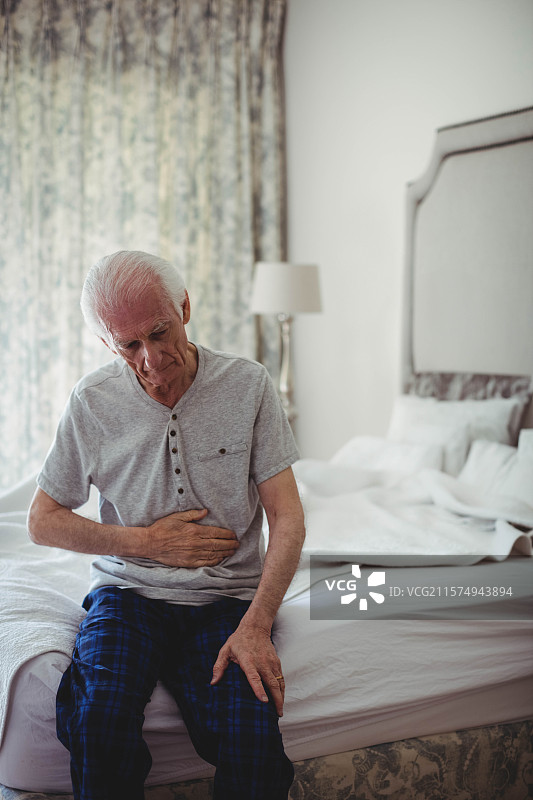 一位老年男子坐在柔和灯光的卧室未整理的床边，窗帘上有花卉图案，旁边有一盏台灯。图片素材
