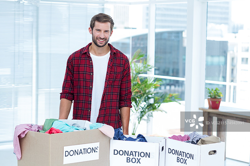 三十多岁的男性志愿者站在社区中心房间里，身后是装满衣物的捐赠箱。图片素材