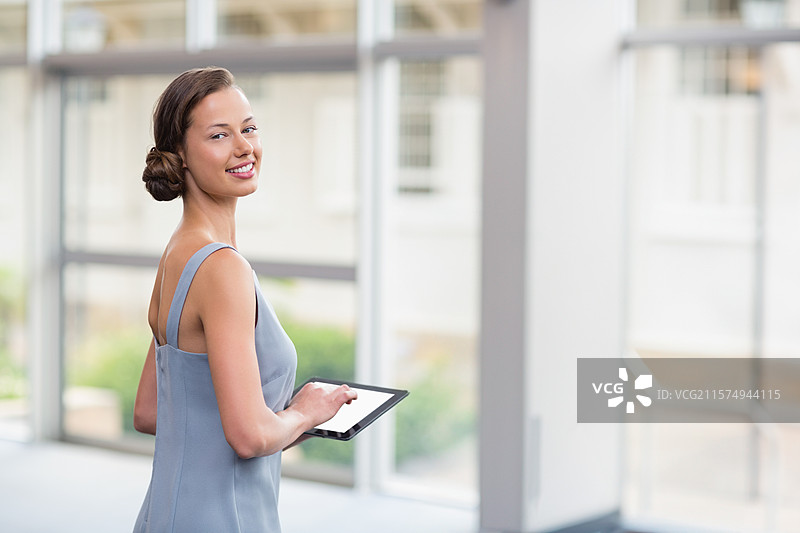 穿着蓝色连衣裙的女性专业人士在办公室走廊中回头看，手持平板电脑，留有空白空间。图片素材