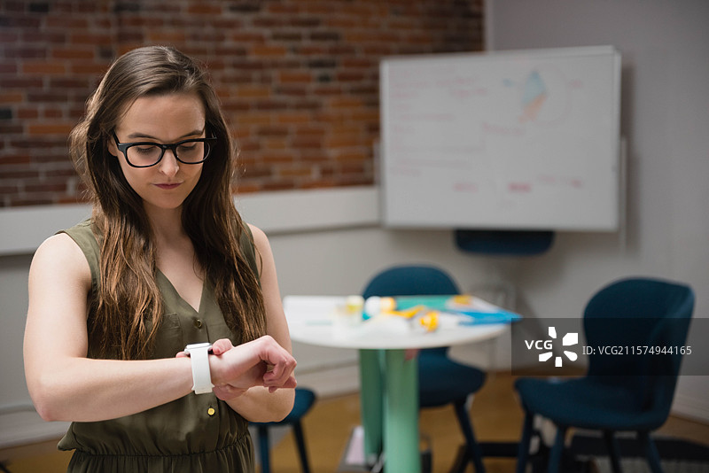 穿着橄榄色连衣裙的女性在办公室查看手腕上的智能手表，背景有白板和椅子，留有空白空间。图片素材