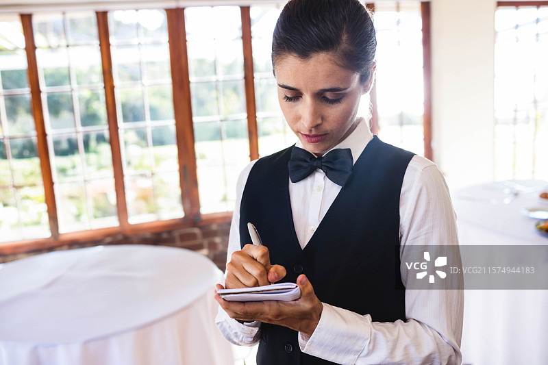 穿着制服的女服务员在宴会厅的白色桌子旁用记事本写字，桌上有玻璃器皿，留有空白空间。图片素材