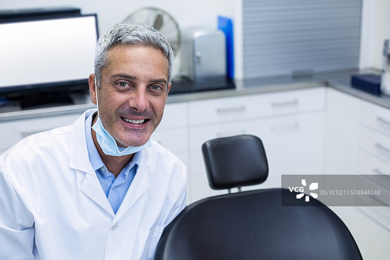 男牙医靠在黑色牙科椅上，诊室内有显示器和摇头风扇，留有空白空间。图片素材