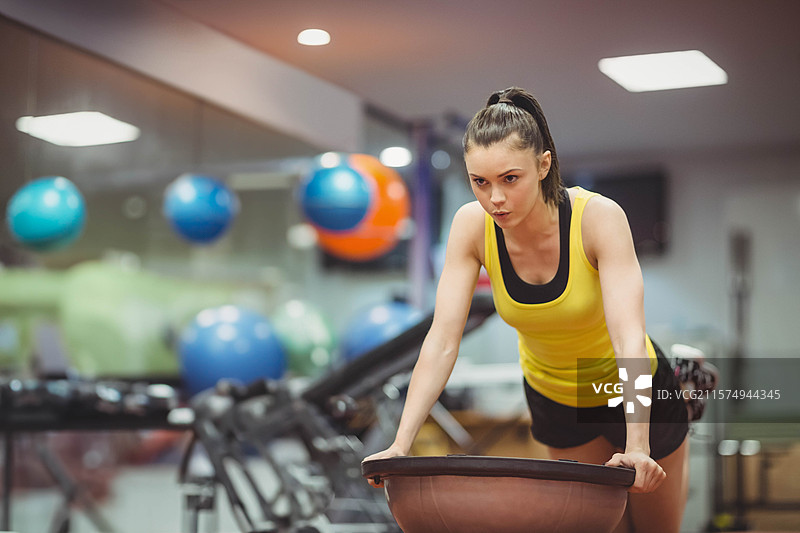 女性在健身房的BOSU平衡训练器上向前倾，旁边是墙壁上挂着的稳定球，留有空白空间。图片素材