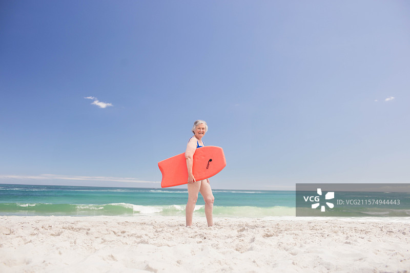 一位老年女性站在白色沙滩上，手持橙色冲浪板，望向碧蓝的海洋。图片素材