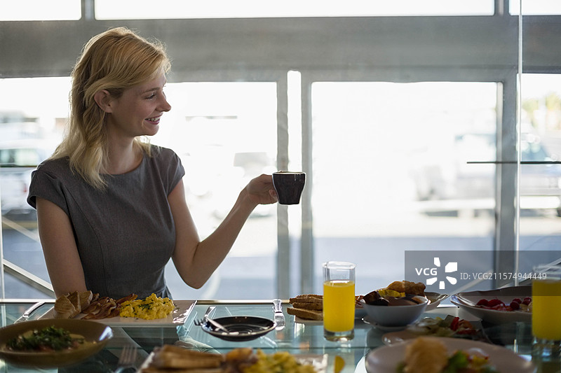 女人坐在玻璃桌咖啡馆里，手里拿着咖啡杯，早餐菜肴一览无余。图片素材