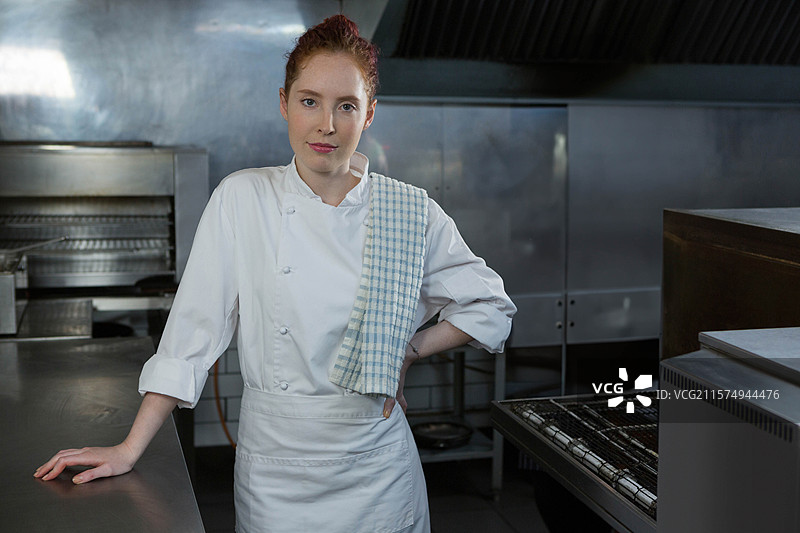 女厨师穿着厨师外套和格子毛巾，靠在厨房的台面上，旁边是烤箱。图片素材