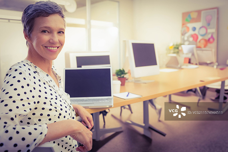 微笑的女性穿着白色波点衬衫，坐在办公室里，旁边有一台关闭的笔记本电脑，留有空白空间。图片素材