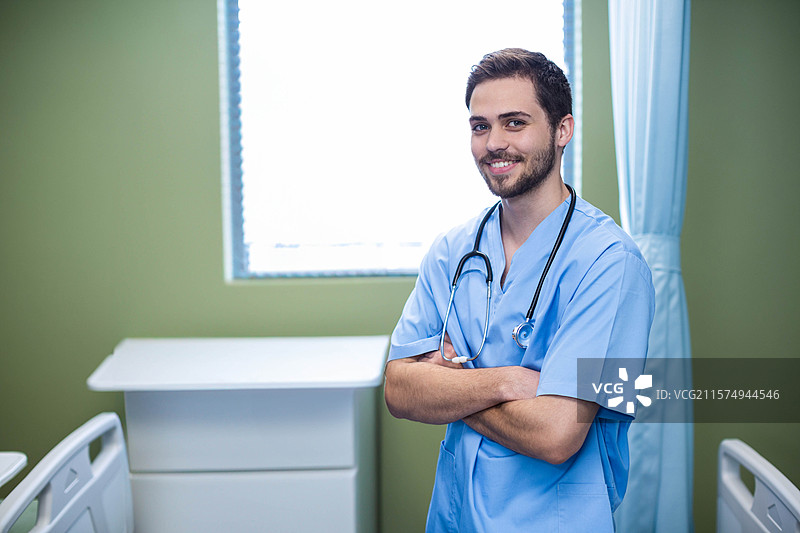 穿着蓝色手术服和听诊器的男医生双臂交叉站在医院病房中，留有空白空间。图片素材