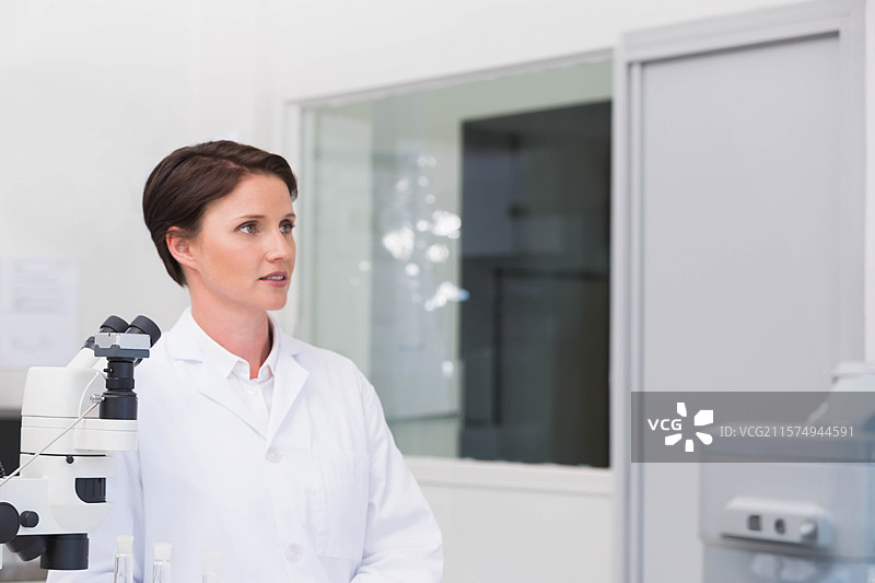 穿着实验室外套的女性科学家在临床实验室中操作显微镜，旁边是诊断分析仪。图片素材