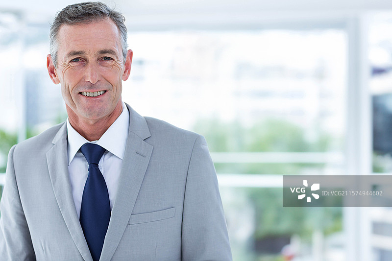 穿着灰色西装的老年男子站在办公室的大窗户旁，微笑着看向镜头。图片素材
