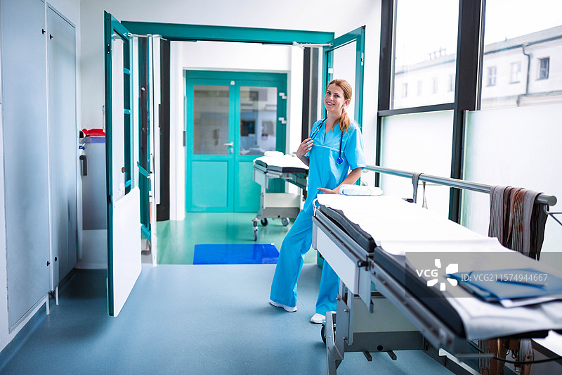 穿着护士服的女性护士站在医院走廊旁边的轮椅担架旁，手里拿着听诊器。图片素材