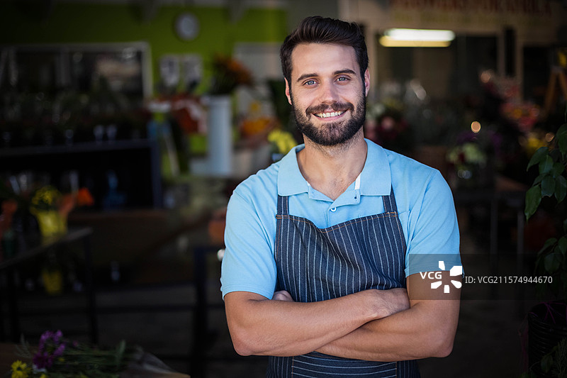 微笑的胡子男人在花店里交叉双臂，穿着条纹围裙，身边是花瓶和植物。图片素材