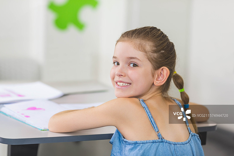 女孩学生坐在灰色桌子旁，翻阅粉色心形作业本，教室内有空白空间。图片素材