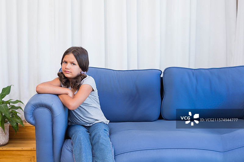女童坐在客厅的蓝色沙发上，旁边有盆栽植物和白色窗帘，留有空白空间。图片素材