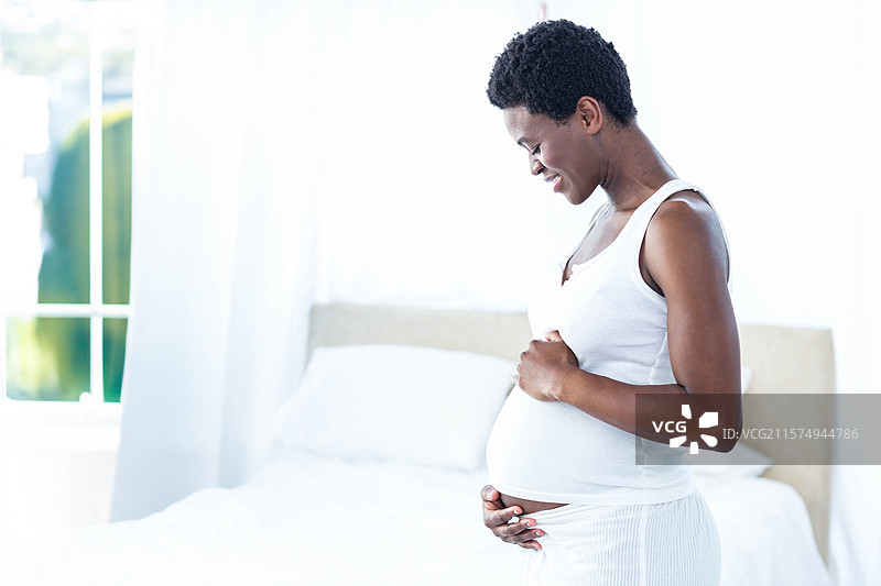 非裔美国女性站在卧室的床边，手放在肚子上，窗帘挂在窗户上，有空白空间。图片素材