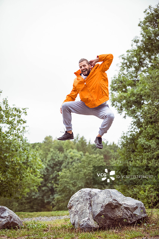 男性穿着亮橙色风衣从大岩石上跳下，位于森林空地的碎石小径上。图片素材