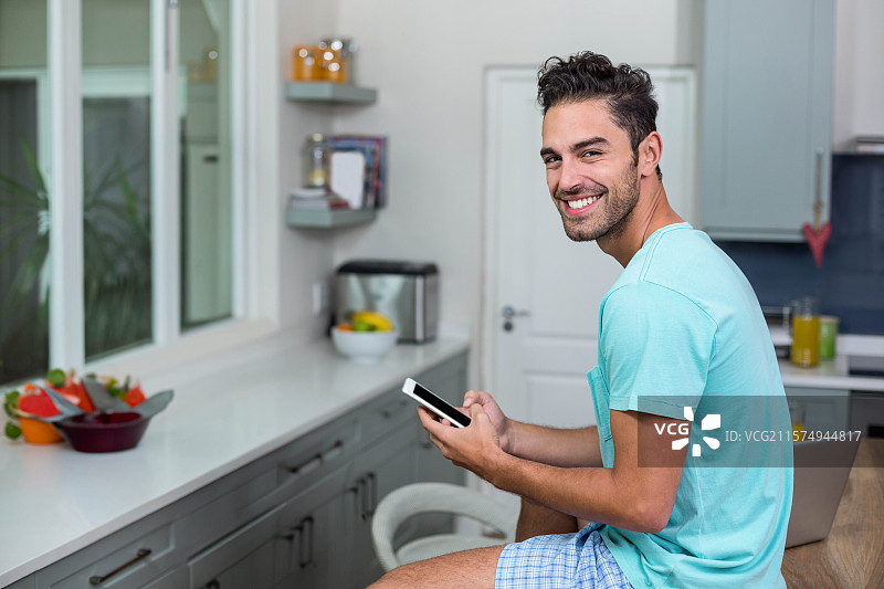 男人坐在厨房台面上的凳子上，手里拿着智能手机和笔记本电脑，旁边有一个水果碗，留有空白空间。图片素材