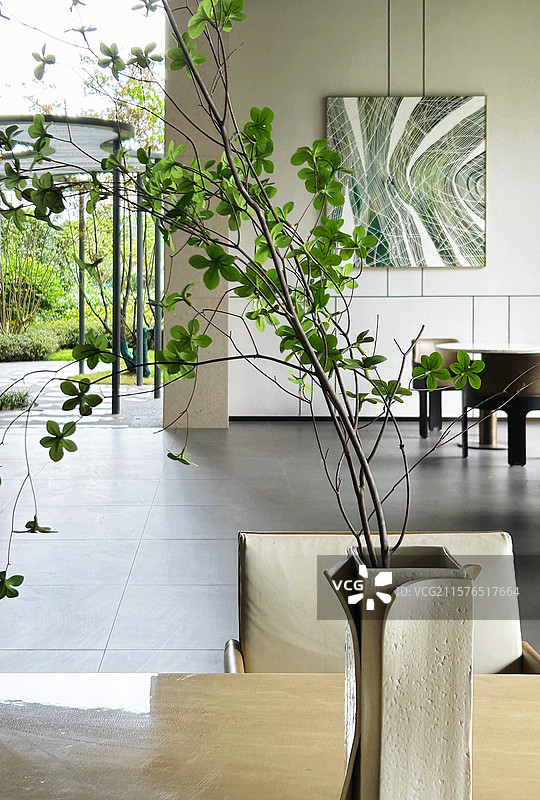 室内浅米色桌上，独特花瓶插着绿枝，背景墙挂艺术画，庭外绿意盎然 。图片素材