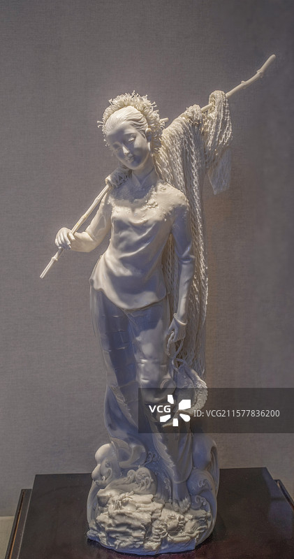 浅色背景前一个右手握着扛在右肩上挂着渔网竹竿左手拎着两只捕获海鱼双脚踏着海浪春风满面归来渔家少女瓷像图片素材