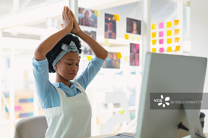 女性在办公桌前冥想时伸展双臂，桌上有电脑、咖啡杯和便利贴。图片素材