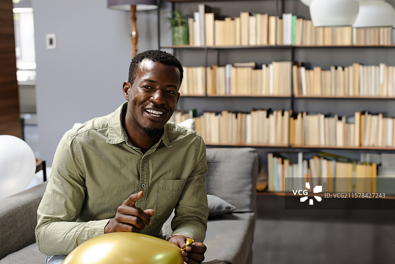 中年非裔美国男子坐在灰色沙发上，为金色气球系上丝带。图片素材