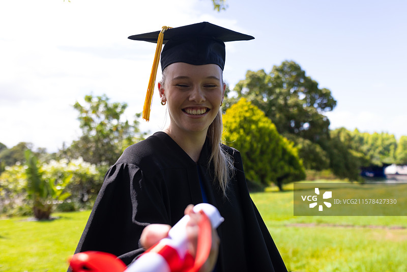 穿着学位袍和带流苏的学士帽的女性毕业生在草坪上递交带红色丝带的毕业证书卷轴图片素材