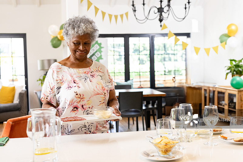 非裔美国老年女性在家中手持装有肉类和奶酪的托盘，周围有气球。图片素材