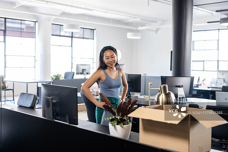 亚洲华裔女性在开放式办公室的桌子旁拆包，桌上有显示器、植物和箱子。图片素材