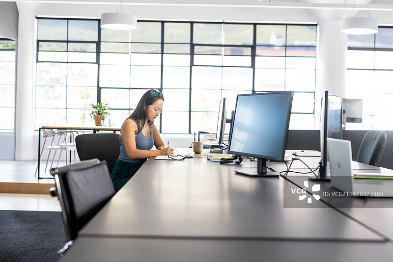 亚洲女性在开放式办公室的桌子上写笔记，旁边有笔记本电脑和咖啡杯，留有空白空间图片素材