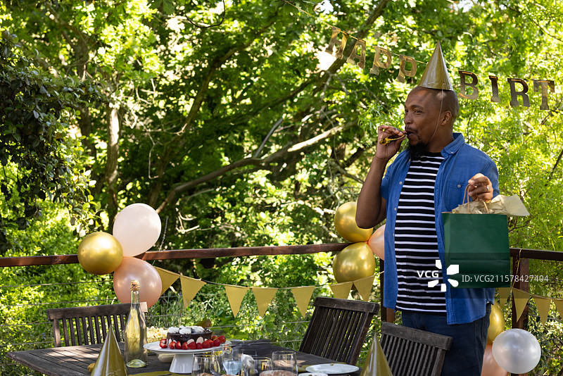 一位非裔美国男性站在木甲板上，手持绿色礼品袋，吹着派对喇叭，留有空白空间。图片素材