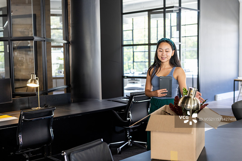 亚洲女性在办公室拆箱，手持平板电脑，取出黄铜灯和仿真植物，留有空白空间。图片素材