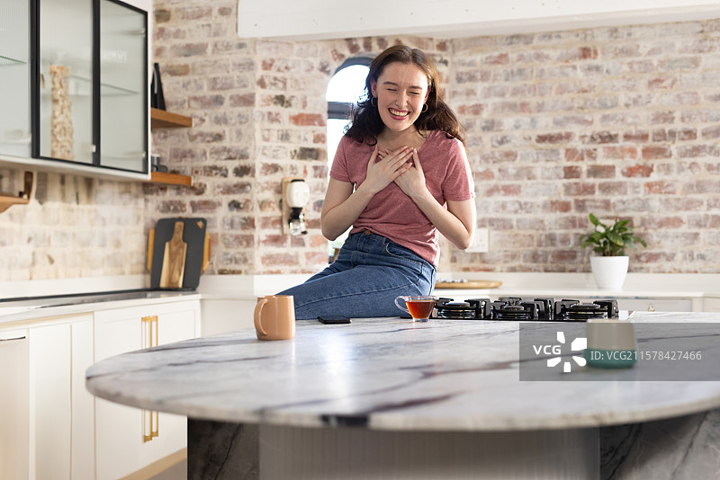 戴着耳罩式耳机的女人在厨房的石材岛上笑着，旁边有几个杯子，留有空白空间。图片素材