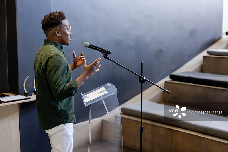 男性演讲者在讲座厅的亚克力讲台前用手势示意，麦克风和纸张靠近奖杯架。图片素材