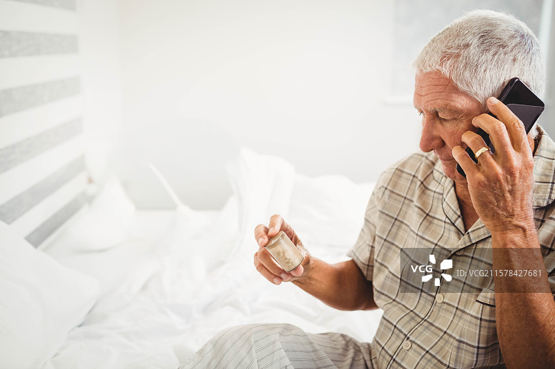 老年男子在条纹床头板的床上，晨光中手持智能手机并检查药瓶。图片素材