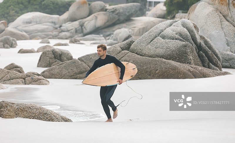 穿着黑色潜水服的成年男性在白色沙滩上走，手里提着带有绳索的木制冲浪板。图片素材