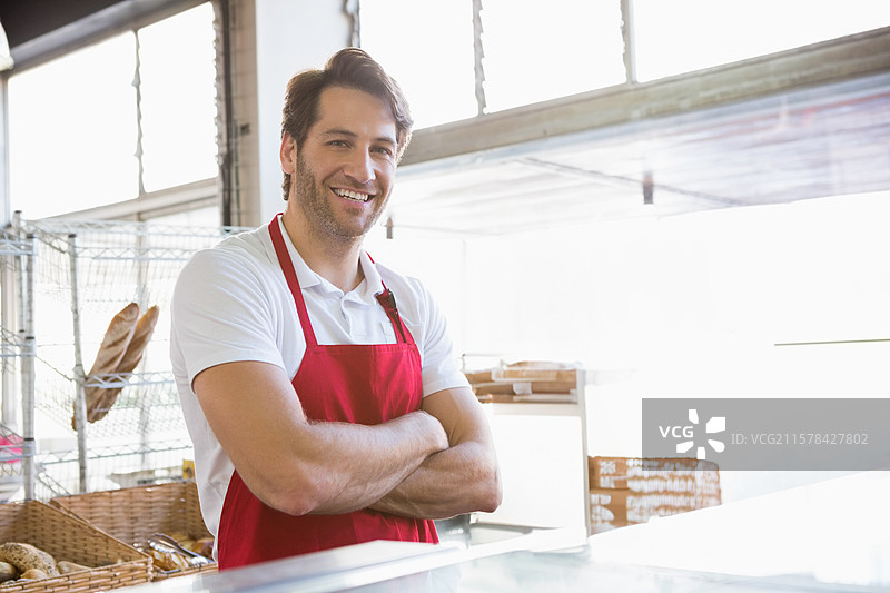 中年男子穿着红色围裙，在面包店的玻璃柜台后工作，旁边有铁丝面包架。图片素材