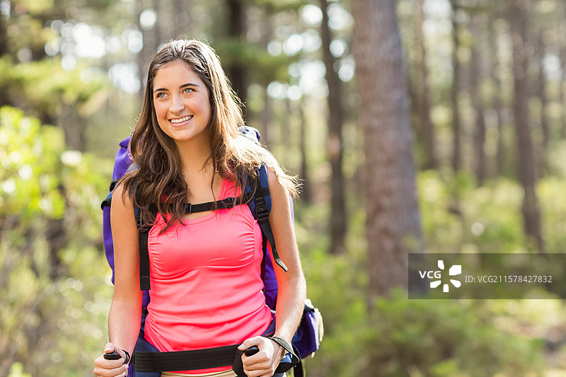 女性徒步旅行者在阳光明媚的松树小径上行走，背着紫色背包，手持登山杖，手腕上戴着手表。图片素材