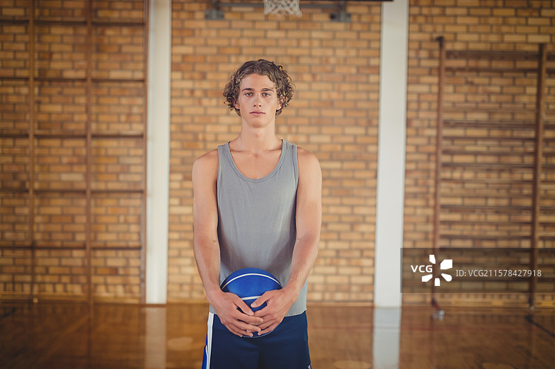 穿着灰色衬衫和短裤的少年在健身房的篮筐下，手里拿着蓝白色篮球，旁边有梯子。图片素材
