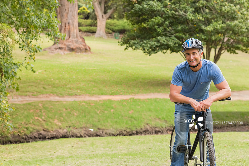 男人靠在黑色自行车上，戴着多彩头盔，站在公园的泥土步道旁，留有空白空间。图片素材