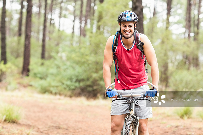 男子骑自行车穿过松树林，戴着头盔，穿着红色背心，戴着蓝色手套，背着背包。图片素材