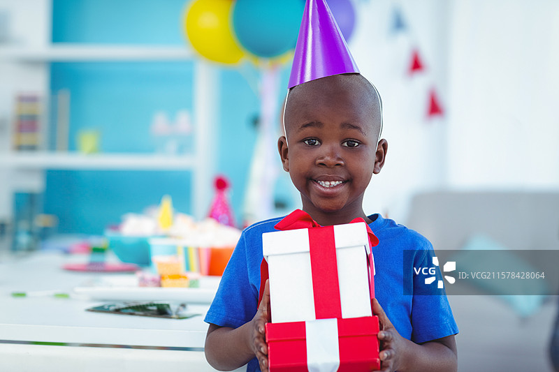 男孩站着，手里拿着礼物盒，客厅里装饰着气球、彩带和派对用品。图片素材