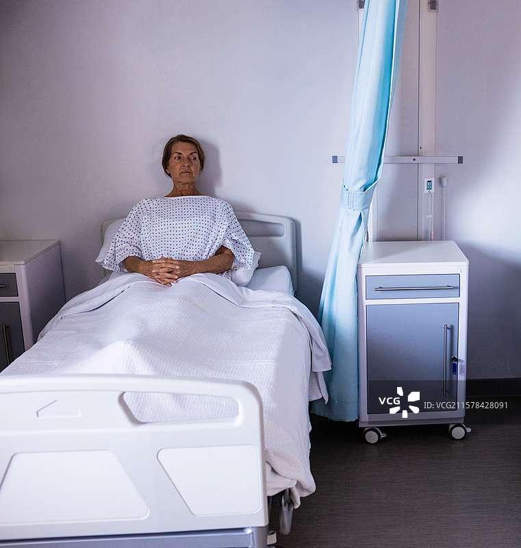 医院病人坐在床上，穿着印花病号服，身边有医疗推车和帘子。图片素材