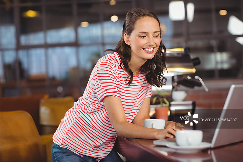 笔记本电脑放在咖啡风格工作区的高木质台面上，旁边有两个咖啡杯和一盆植物。图片素材