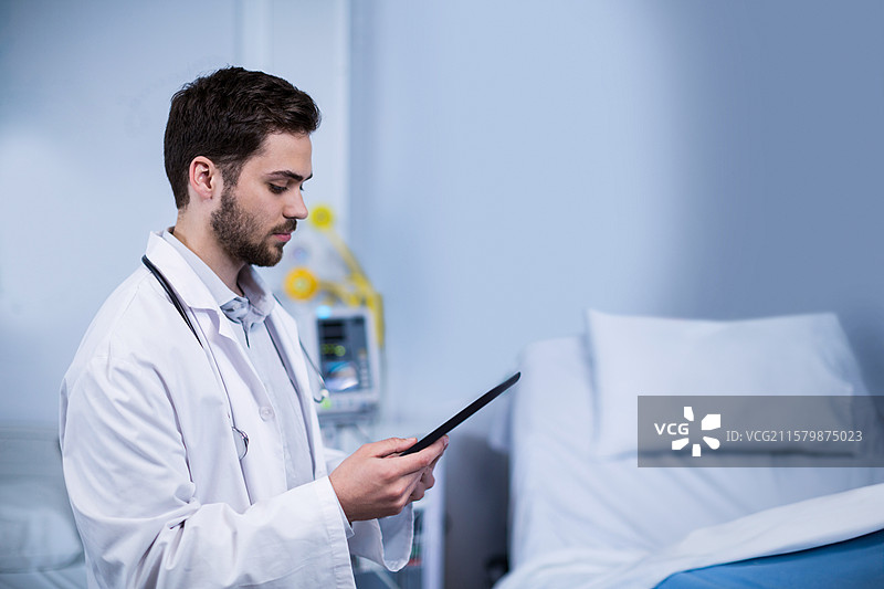 男医生在床边查看平板电脑，身旁有听诊器、蓝色毛毯和监视器，留白空间。图片素材
