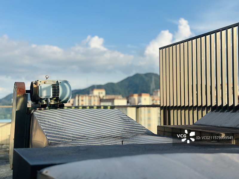 天台一隅，机器、光影与远山共绘日常。蓝天缀着云朵，山峦在城市建筑后沉默，明暗条纹里，藏着喧嚣中的宁静图片素材