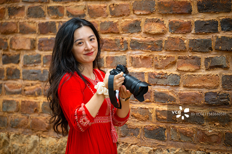 红衣女子手持相机站在砖墙前图片素材
