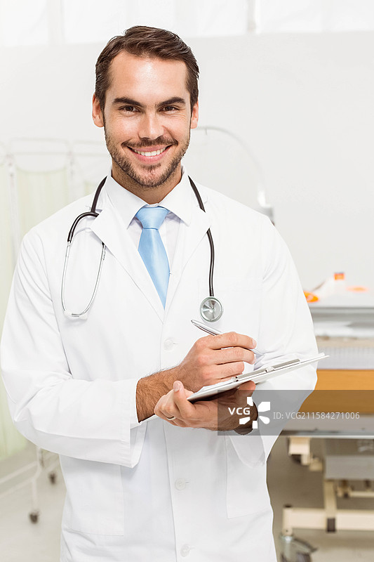 穿着白大褂和蓝色领带的男性医生在检查室里手持夹板，脖子上挂着听诊器。图片素材
